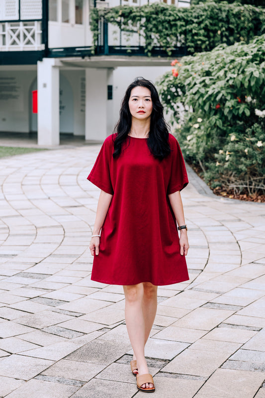 Woman in a knee length burgundy linen dress standing on a paved walkway with greenery in the background
