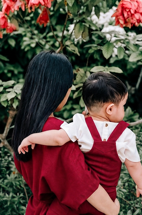 A woman in a burgundy dress carries a toddler wearing matching burgundy linen dungaree.