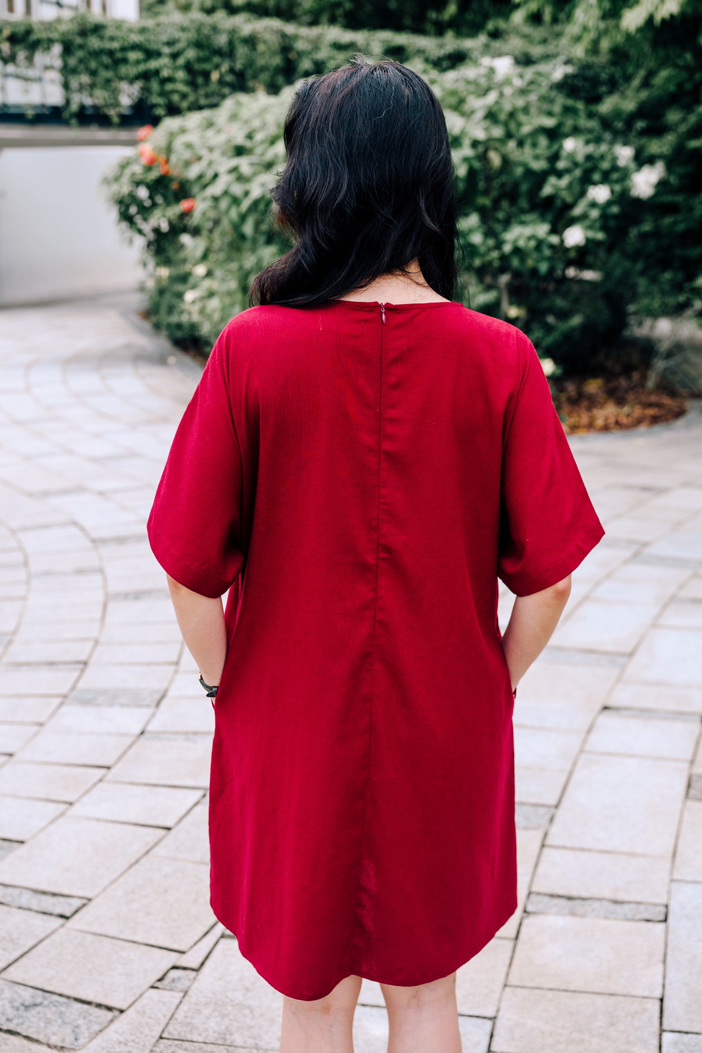 Back view of woman wearing a knee length burgundy linen dress standing on a paved walkway