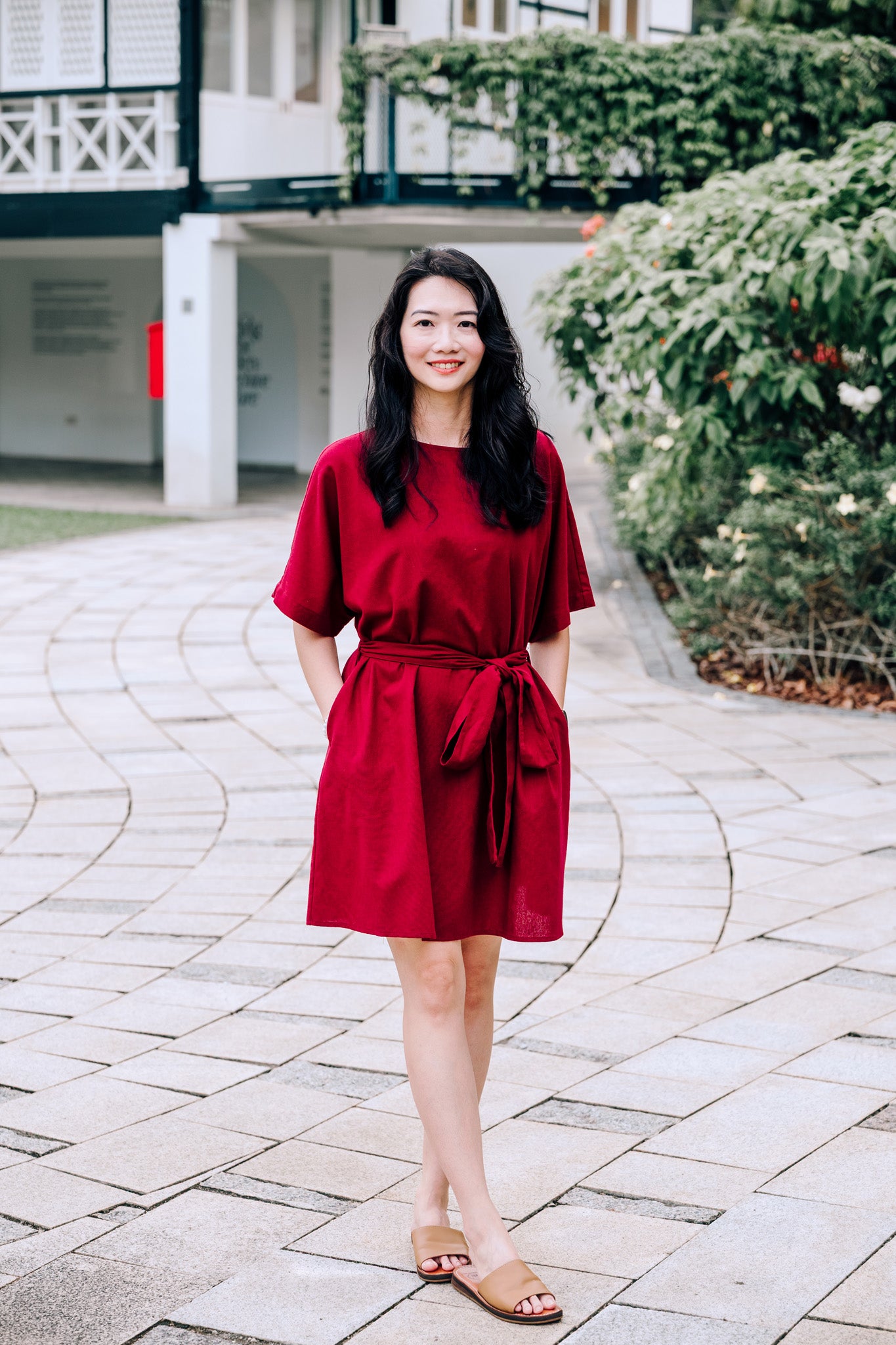 Woman in a knee length burgundy linen dress standing on a paved walkway with greenery in the background
