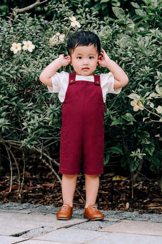 A toddler wearing burgundy linen overalls over a white short-sleeve shirt and brown shoes.
