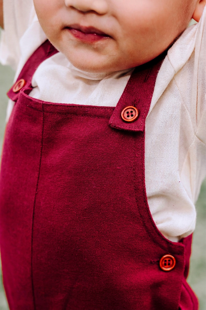 Close-up of boy wearing burgundy linen overalls over a sand beige linen t-shirt. 